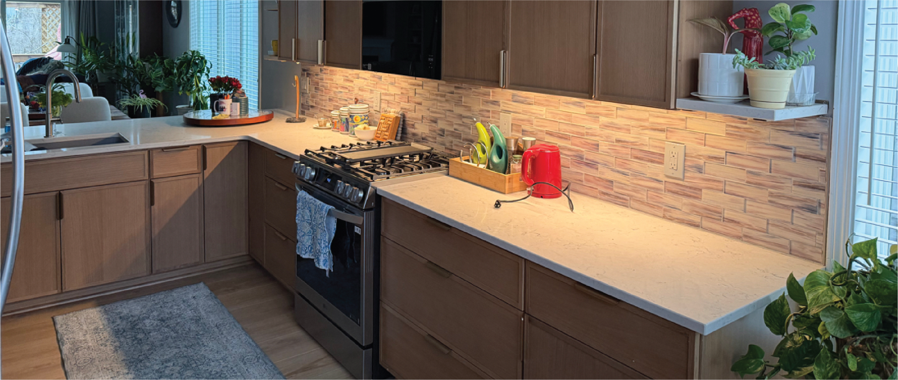 Modern kitchen with light wood cabinets, white countertops, an under-cabinet lighted tiled backsplash, a stainless steel gas range, and various countertop plants and accessories.