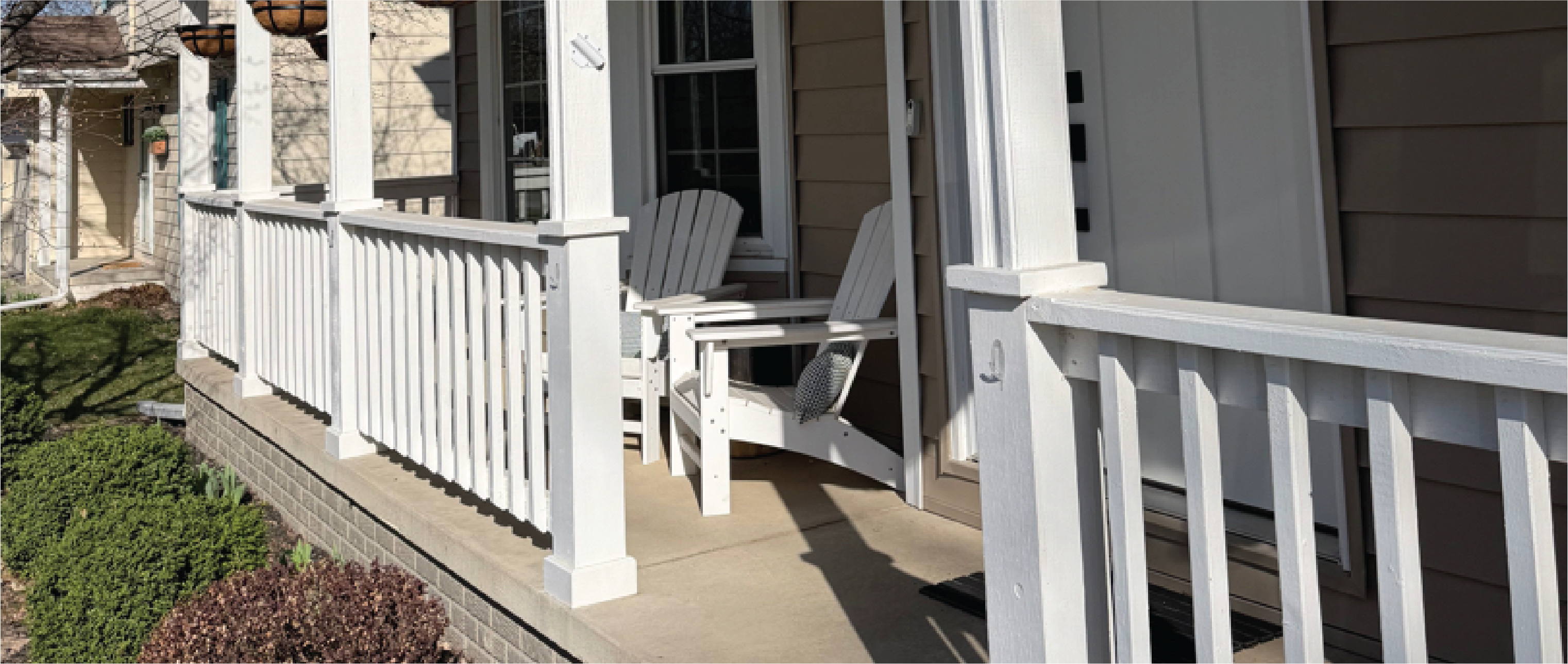 A cozy front porch with tan siding, crisp white railings, and matching support pillars. Two white Adirondack chairs with patterned pillows face outward, creating an inviting seating area. Green manicured bushes line the brick foundation below.