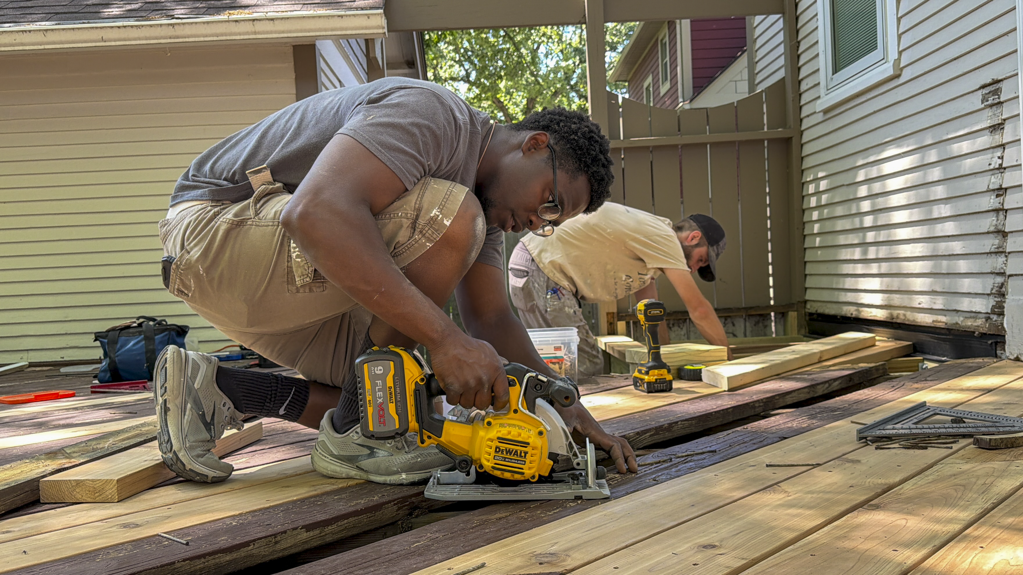 A contractor uses a DeWalt circular saw to cut deck boards during a backyard deck repair project, with tools and another worker measuring boards on a partially built wooden deck.