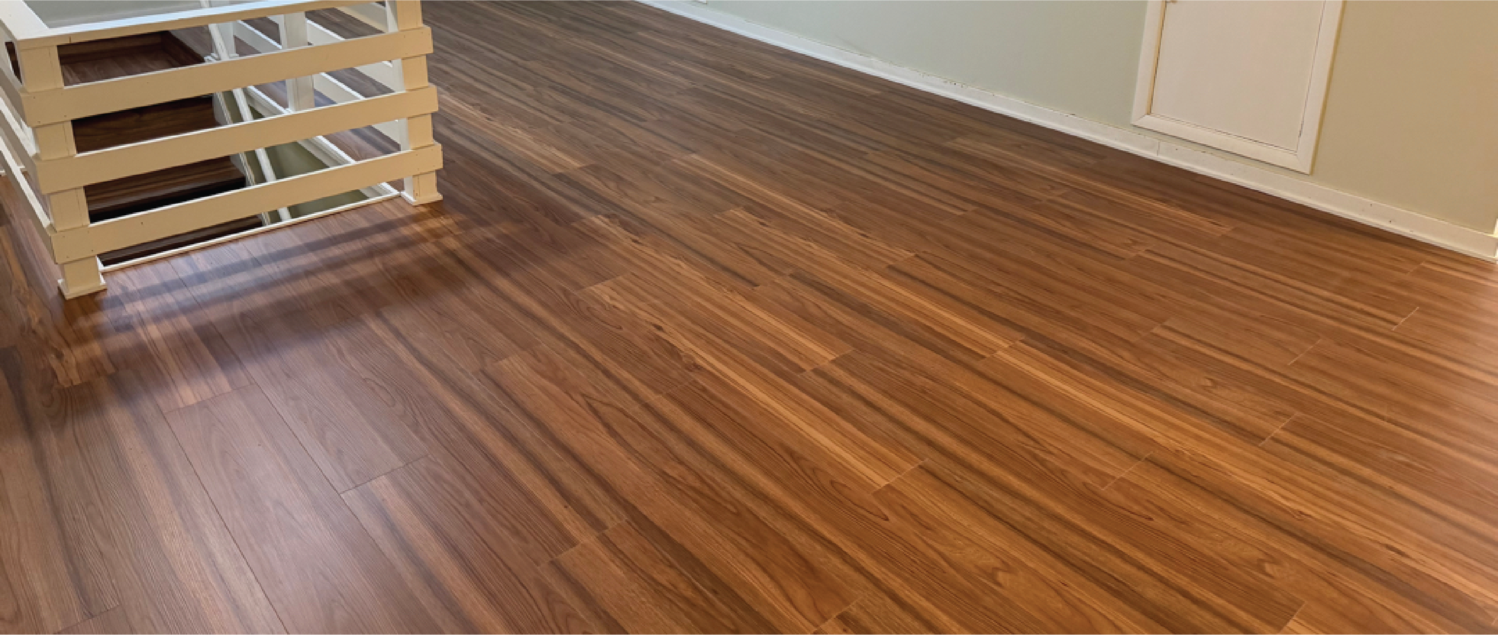 A wide shot of a newly installed or polished medium-brown wood laminate or hardwood floor. In the upper-left corner, a white wooden safety railing frames the top of a staircase leading down. The walls are a light neutral tone with white baseboards and a closed white door visible in the upper right.